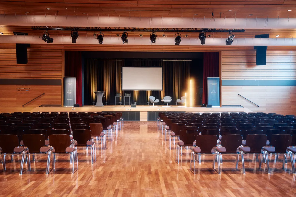 Leerer Saal mit Holzwänden, Stuhlreihen und beleuchteter Bühne